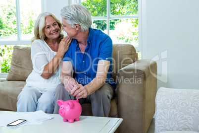 Cheerful senior couple putting coin in piggi bank at home