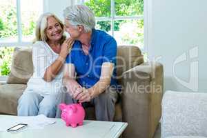 Cheerful senior couple putting coin in piggi bank at home