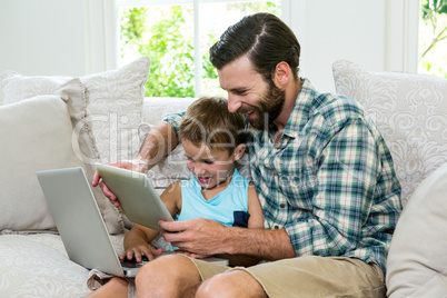 Playful son and father with laptop and digital tablet on sofa