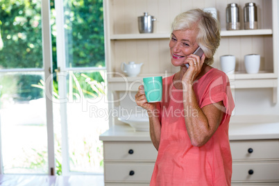 Happy senior woman talking on phone while having coffee in kitch