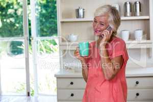 Happy senior woman talking on phone while having coffee in kitch