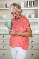 Senior woman using mobile phone while having coffee in kitchen