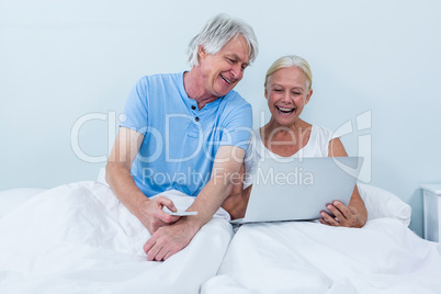 Happy retired couple using laptop while sitting on bed