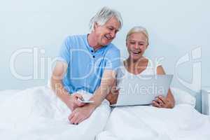 Happy retired couple using laptop while sitting on bed