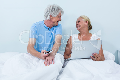 Cheerful retired couple using laptop while sitting on bed