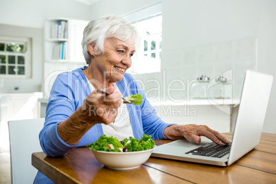 Senior woman eating salad while using laptop at table