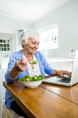 Smiling senior woman eating salad while using laptop at table