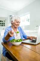 Smiling senior woman eating salad while using laptop at table