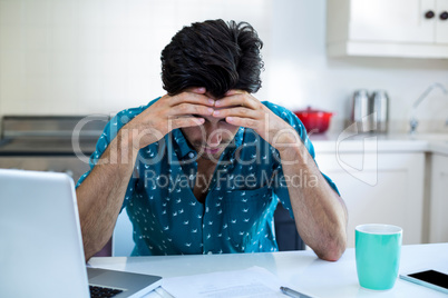 Tensed man sitting with bills and laptop in kitchen