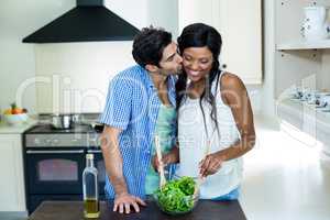 Man kissing a woman while mixing a salad in kitchen