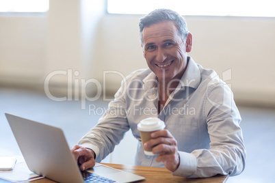 Businessman holding disposable coffee cup and using laptop