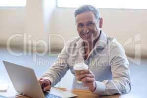 Businessman holding disposable coffee cup and using laptop