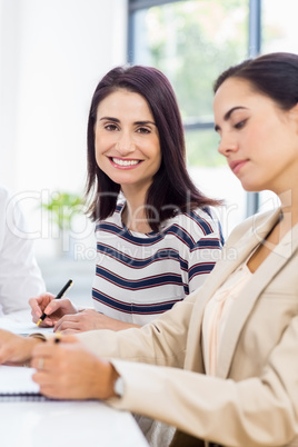 A businesswoman is posing and smiling and another is concentrati