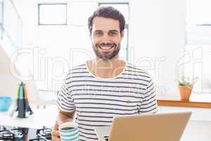 Portrait of happy man holding coffee cup and laptop in kitchen