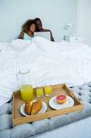 Young couple using a laptop with breakfast tray ready on bed
