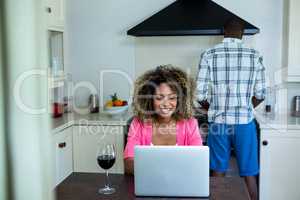 Woman using laptop while man cooking food in background