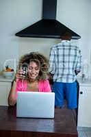 Woman using laptop and having wine while man cooking food in bac