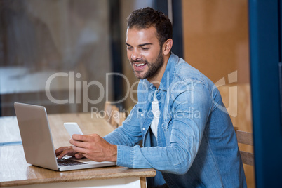 Man text messaging on phone while sitting on desk with laptop