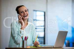 Woman talking on mobile phone while sitting on desk with laptop