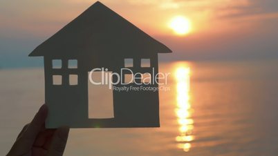 Hand holding cut paper house against of sea at sunset
