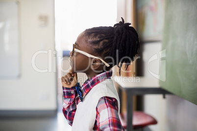 Thoughtful schoolgirl standing with hand on chin in classroom