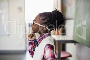 Thoughtful schoolgirl standing with hand on chin in classroom