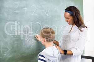 Smiling teacher assisting boy in doing addition on chalkboard