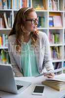 Thoughtful teacher using digital tablet with laptop and phone on table in library