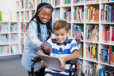 Happy schoolgirl standing with schoolboy on wheelchair using digital tablet