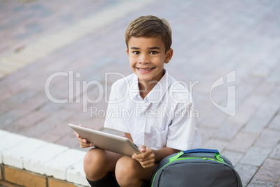 Happy schoolboy sitting in campus and using digital tablet