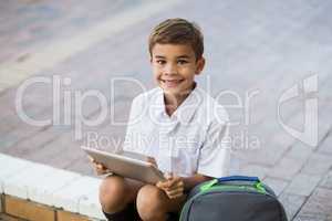 Happy schoolboy sitting in campus and using digital tablet
