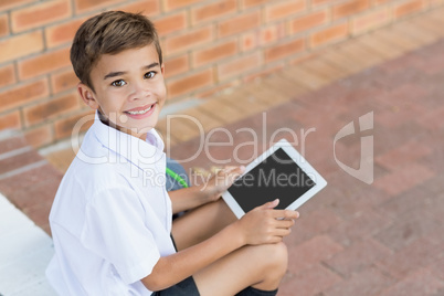 Happy schoolboy sitting in corridor and using digital tablet