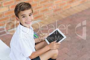 Happy schoolboy sitting in corridor and using digital tablet