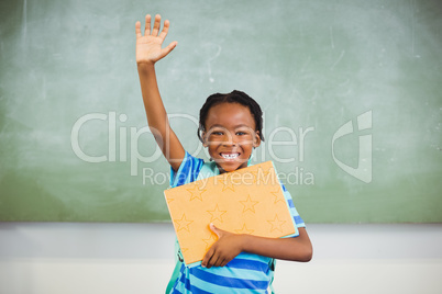 Happy schoolboy raising his hand and holding books in class room
