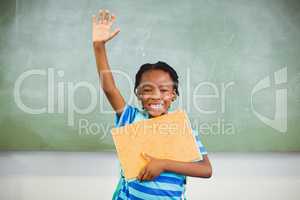 Happy schoolboy raising his hand and holding books in class room