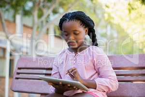 Schoolgirl sitting on bench and using digital tablet in campus