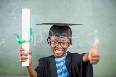 Schoolboy in mortar board holding certificate and showing thumbs up in classroom
