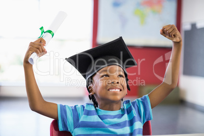 Excited schoolboy in mortar board holding certificate in classroom
