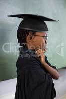 Thoughtful schoolboy wearing graduation gown in classroom