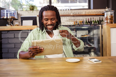 A man is drinking a cup of coffee while is looking his tablet