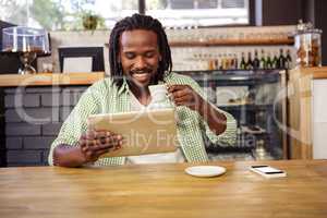 A man is drinking a cup of coffee while is looking his tablet