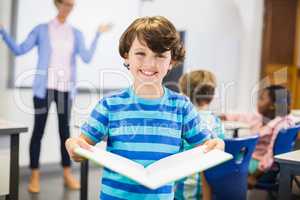 Portrait of schoolboy standing with book in classroom