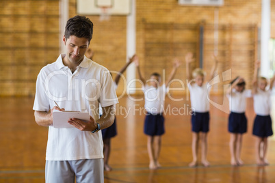 Sports teacher writing notes on clipboard while students exercis