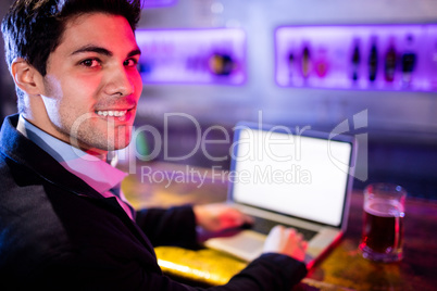 Smiling man using laptop with glass of beer on table at bar coun