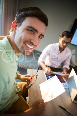 Man smiling at camera while his colleague reading document