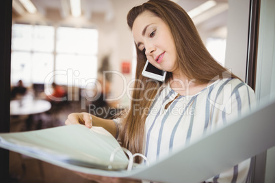 Young businesswoman holding files while using mobile phone in of