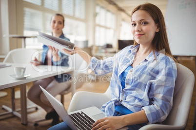 Businesswoman giving file to colleague while working on laptop a