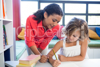 Teacher assisting girl writing on notebook in library