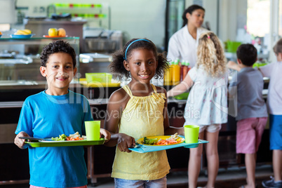 Happy schoolchildren holding food tray in canteen