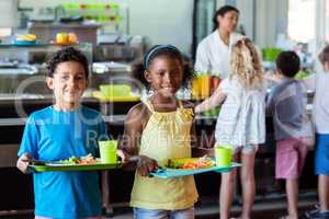 Happy schoolchildren holding food tray in canteen